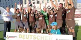 Hinter einem, von zwei Kindern gehaltenen Schild mit der Aufschrift "Deutscher Meister" steht das FC St. Pauli-Team in braunen Trikots und jubelt in die Kamera. Hippo hält die Meisterschale