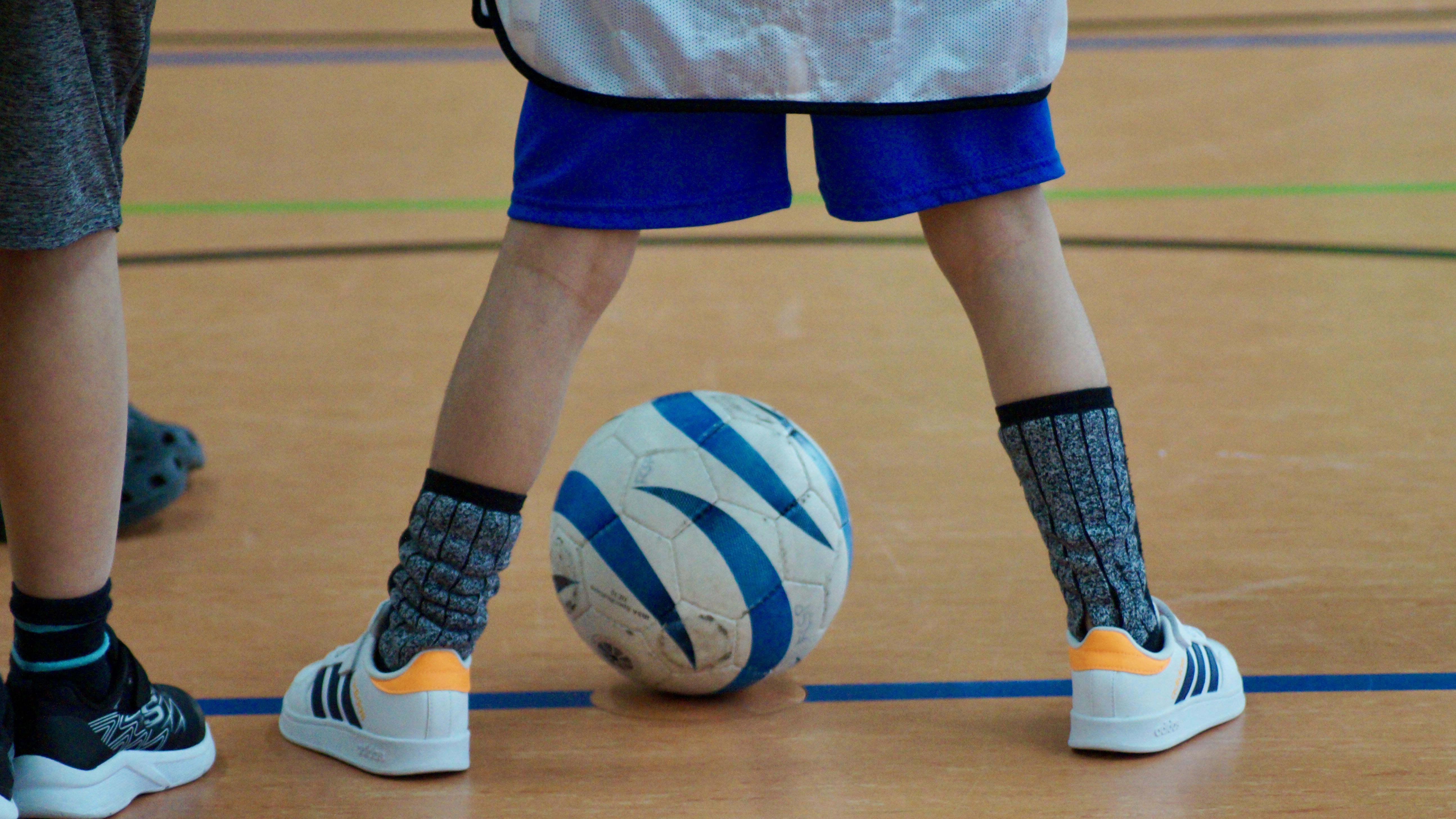 Ein Blindenfußball ist durch zwei Kinderbeine auf dem Hallenboden zu sehen