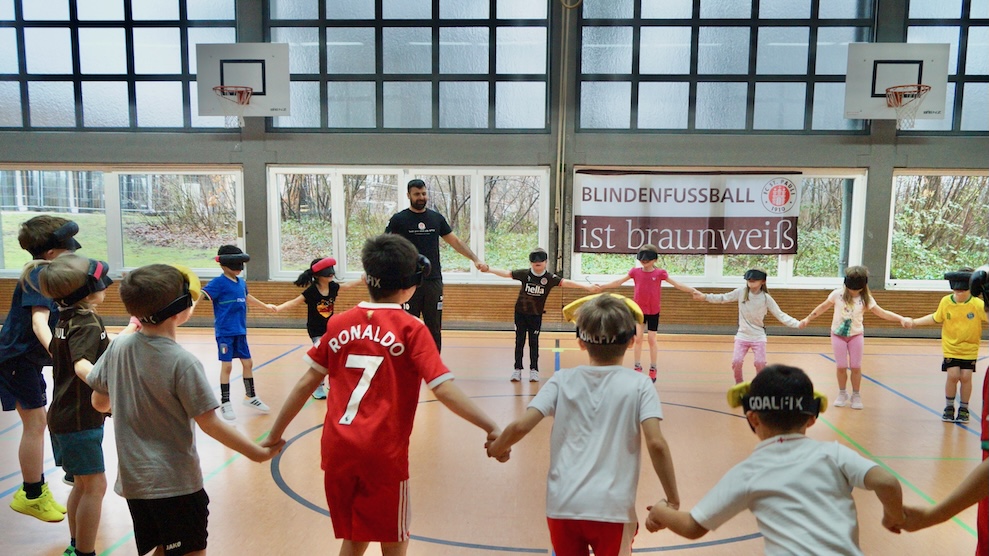 Serdal leitet in einer halle eine Gruppe Kinder an, die sich alle im Kreis an den Händen halten