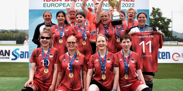 Teamfoto vor der Sponsorenwand mit Pokal. Alle jubeln und lachen